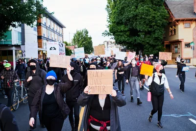 Masked protesters fill the asphalt streets of Portland's Kerns neighborhood during a Black Lives Matter demonstration in the aftermath of George Floyd's murder. The diverse crowd moves through the tree-lined residential area near East Burnside Street, carrying handmade signs with messages of justice and solidarity against a backdrop of characteristic Pacific Northwest architecture. The late afternoon light casts long shadows across the pavement as the peaceful march progresses through this historic Portland community.