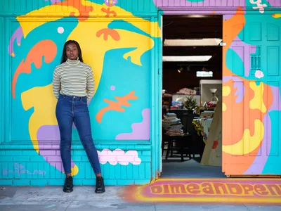 Naomi Likayi stands confidently against her newly completed mural in Portland's Alberta Arts District, where swirling abstract forms in electric turquoise, sunshine yellow, coral orange, and lavender purple transform the storefront facade. The young Black artist wears a cream striped turtleneck and dark jeans, her relaxed pose contrasting with the explosive energy of her artwork that flows around the entrance to what appears to be a community bookstore. Natural daylight illuminates both the artist and her bold street art, creating a powerful portrait of creative accomplishment in this gentrifying Northeast Portland neighborhood.