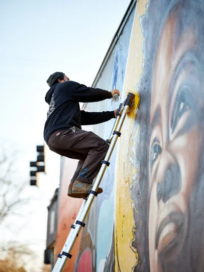 Travis Fields, known as campographic, balances on a telescoping ladder against a weathered wall, applying vibrant yellow paint to his Amanda Gorman mural at Northeast 15th and Killingsworth in Portland's Alberta district. The artist works with focused concentration under overcast skies, his dark hoodie and beanie contrasting against the emerging colors of the tribute piece. Urban residential buildings blur softly in the background, creating depth while emphasizing the intimate scale of this community art project.