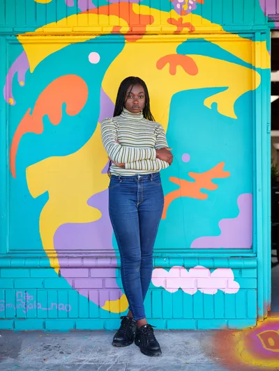 Naomi Likayi stands confidently with crossed arms before her completed mural in Portland's Alberta Arts District. The artist wears a striped turtleneck and dark jeans, positioned against an explosion of turquoise, yellow, orange, and purple abstract forms that dance across the brick wall behind her. Natural daylight illuminates both subject and artwork, creating a harmonious dialogue between creator and creation in this Northeast Portland neighborhood known for its thriving street art scene.
