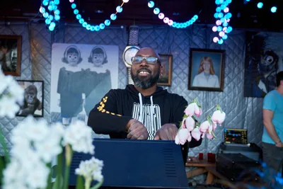 DJ Cee White, wearing reflective sunglasses and a black hooded sweatshirt, commands the decks at White Owl Social Club's intimate venue space. Cascading string lights create ethereal bokeh patterns above vintage portrait photographs that line the diamond-quilted walls, while pink tulips in the foreground add unexpected softness to this Portland nightlife scene. The atmospheric lighting and eclectic gallery setting transform the day party into a moody sonic sanctuary.