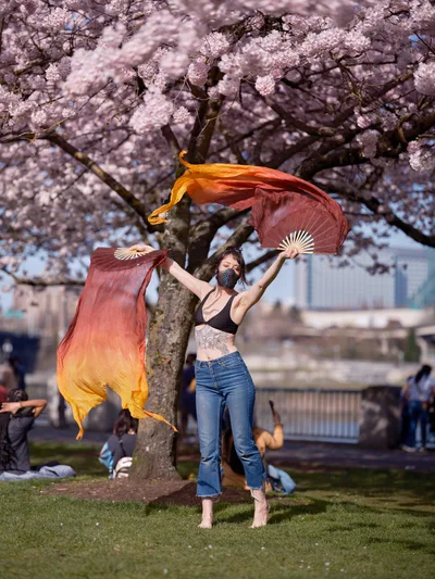 A performer with flowing silk fans dances gracefully beneath a canopy of pale pink cherry blossoms at Tom McCall Waterfront Park in Portland, Oregon. The dancer, wearing a black crop top, distressed jeans, and face mask, creates fluid motion with gradient orange-to-burgundy silk fans that catch the soft spring light. Behind her, the historic branches of mature cherry trees frame the scene while parkgoers relax on the grass, creating an intimate moment of artistry against the backdrop of Portland's downtown skyline.