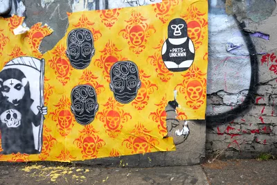 A weathered wheat paste installation covers a cracked concrete wall in Portland's Central Eastside, featuring bold yellow damask wallpaper adorned with black skull imagery and a wrestler-masked figure. The deteriorating urban canvas bears the scars of time, with torn edges and faded stencil work creating a palimpsest of street art layers. Red spray paint bleeds through beneath the paste-up, while a partial Grim Reaper stencil anchors the composition's left edge.