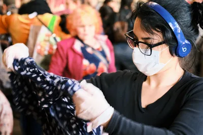 A shopper wearing a white face mask, black-rimmed glasses, and blue Beats wireless headphones examines a patterned garment at a crowded swap meet inside the Crystal Ballroom in Portland, Oregon. She wears a black long-sleeve top and is focused on inspecting the clothing item. A busy crowd of other shoppers is visible in the blurred background.