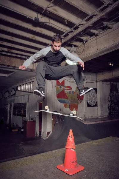 A focused skateboarder executes a precise ollie over a weathered orange traffic cone in the atmospheric basement of Commonwealth Skateboarding in Portland's Buckman neighborhood. The industrial space features exposed ceiling joists, graffitied concrete walls, and a makeshift skate bowl, while warm overhead lighting illuminates the concentrated athlete mid-trick. The gritty urban interior, with its painted murals and raw concrete surfaces, captures the authentic underground skateboarding culture of Southeast Portland.