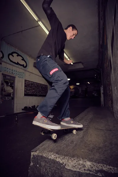 A focused skateboarder in gray work pants and casual sneakers glides through the interior of Commonwealth Skateboarding in Portland's Buckman neighborhood. The atmospheric scene captures him mid-ride beneath harsh fluorescent lighting, with vintage skateboard graphics and weathered concrete steps creating an authentic underground skate culture backdrop. His concentrated expression and fluid motion through this iconic Portland skate shop epitomize the dedication and flow that defines street skating.