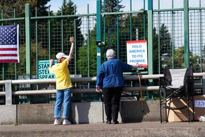 Two protesters position themselves along the North Skidmore overpass above Interstate 5 in Portland's Humboldt neighborhood, displaying signs calling for civic engagement and a May 1st General Strike. A woman in a bright yellow shirt raises her hand in greeting while an elderly man with a walking cane adjusts a red, white and blue protest banner reading 'Hold America to Its Promise.' The green mesh security fencing and concrete barriers frame their grassroots demonstration against the backdrop of evergreen trees under overcast Pacific Northwest skies.