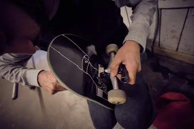 In the intimate workspace of Commonwealth Skateboarding on Southeast 20th Avenue, focused hands adjust the trucks and wheels of a black skateboard deck adorned with linear graphics. The moody lighting casts deep shadows across weathered concrete floors, while the skater's deliberate movements speak to the meticulous craft of board assembly. This behind-the-scenes moment captures the tactile relationship between rider and equipment in Portland's vibrant skate culture.