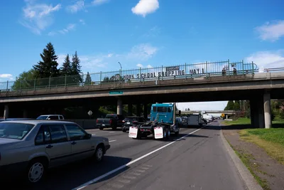 Activists from the Indivisible movement unfurl a protest banner reading 'No Work School Shopping May 1st' across the North Skidmore Street overpass above Interstate 5 in Portland's Humboldt neighborhood. Heavy afternoon traffic flows beneath the concrete bridge as towering evergreens frame the scene under partly cloudy Pacific Northwest skies. The grassroots demonstration calls for a general strike, transforming urban infrastructure into a platform for political messaging.