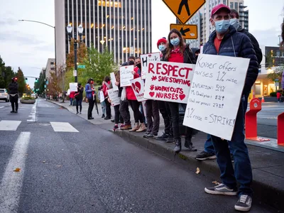 Healthcare workers line the rain-slicked streets of Portland's Lloyd District, their handwritten protest signs catching the amber glow of evening streetlights against towering medical office buildings. Masked demonstrators in scrubs and casual wear hold placards demanding respect, safe staffing levels, and fair compensation, their determined faces reflecting the gravity of their cause. The wet pavement mirrors the urban canyon of glass and steel that houses Kaiser Permanente's regional headquarters, creating a stark juxtaposition between corporate power and grassroots advocacy. The photographer captures this moment of collective action with intimate street-level perspective, emphasizing the human scale of the protest against the institutional backdrop.
