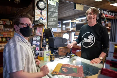Two music enthusiasts connect across the glass counter at Music Millennium during Record Store Day 2022, their shared passion for vinyl evident in the warm exchange. The masked customer in plaid shirt leans forward while the店owner in black long-sleeve shirt with circular logo stands behind the register, surrounded by the eclectic chaos of badges, policy signs, and musical memorabilia that defines this iconic Portland record shop. Fluorescent lighting bathes the scene in the authentic glow of independent retail culture, capturing a moment where analog music brings people together.