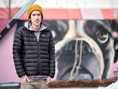 Muralist Weti stands confidently before his vibrant street art creation on the exterior wall of Clay Creative Building in Southeast Portland's Hosford-Abernethy neighborhood. Wrapped in a black down jacket and golden knit beanie against the Pacific Northwest chill, the tattooed artist poses with quiet satisfaction beside his large-scale work featuring bold geometric forms and watchful eyes rendered in pink, black, and cream tones. The urban portrait captures both the creator and creation in sharp focus, while the colorful mural provides a dynamic backdrop that speaks to Portland's thriving street art culture.