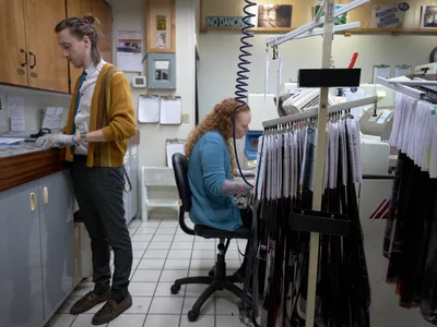 In the intimate workspace of Blue Moon Camera & Machine on North Lombard Street, two technicians pursue the meticulous craft of analog photography development. A young man in a golden cardigan examines film strips at a light table while his colleague, wearing a blue sweater, operates specialized processing equipment surrounded by hanging negatives. The fluorescent-lit lab, dense with filing systems and vintage photography notices, captures the enduring artistry of traditional film processing in Portland's Saint Johns neighborhood.