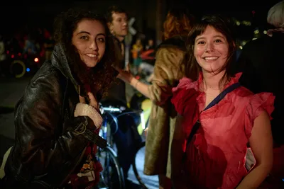 Two young women beam with infectious joy during Portland's Drop Out Prom Ride, their contrasting outfits—one in weathered olive military jacket, another in vibrant coral ruffled dress—creating a striking visual dialogue against the blurred nocturnal revelry. The warm glow of streetlights and scattered bicycle lights illuminate their genuine smiles, capturing the spontaneous intimacy of friendship amid the organized chaos of Pedalpalooza's unconventional celebration in Southeast Portland's Hosford-Abernethy neighborhood. Behind them, fellow participants blur into impressionistic figures, their presence suggesting the communal energy of this beloved alternative prom experience.