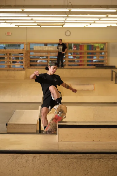 Professional skateboarder Christiana Means demonstrates fluid skateboarding technique as she navigates the curved concrete bowl at Stronger Skatepark in Milwaukie, Oregon. Her focused expression and athletic form are captured mid-maneuver, with warm fluorescent lighting illuminating the indoor facility's wooden observation deck and neutral concrete surfaces. A fellow skater observes from the elevated platform, creating depth in this dynamic action portrait.