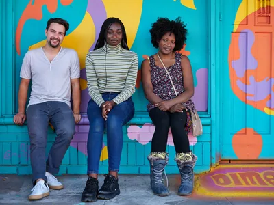 Travis Fields, Naomi Likayi, and the owner of the Dime & Penny sit together on the steps of a vibrant turquoise building adorned with bold abstract murals in Portland's Alberta Arts District. The trio radiates warmth and community spirit against the kaleidoscope of coral, yellow, and purple paint that transforms the urban storefront into a canvas of creative expression. Natural daylight illuminates their relaxed poses and genuine smiles, capturing an intimate moment of friendship and local business camaraderie. The colorful street art backdrop perfectly embodies the artistic energy that defines this Northeast Portland neighborhood.