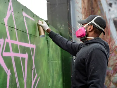 A dedicated street artist wearing a bright pink respirator mask carefully applies green spray paint to a concrete wall, adding bold strokes to an emerging mural. The scene unfolds in the parking lot of Clay Creative in Southeast Portland's Hosford-Abernethy neighborhood, where layers of existing graffiti create a colorful backdrop. Soft daylight illuminates the artist's focused concentration as he extends his arm to reach higher portions of the wall, his black hoodie and cap contrasting against the vibrant pink and green paint.
