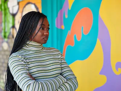 Naomi Likayi stands in quiet contemplation before her newly completed mural on Northeast 15th Avenue in Portland's Alberta district. Her long black braids cascade over a striped turtleneck as she gazes thoughtfully at the explosion of coral, turquoise, and golden hues that dance across the wall behind her. The organic, flowing forms of her street art create a striking contrast against her measured, reflective pose, capturing a moment of artistic satisfaction in this gentrifying corner of Northeast Portland.