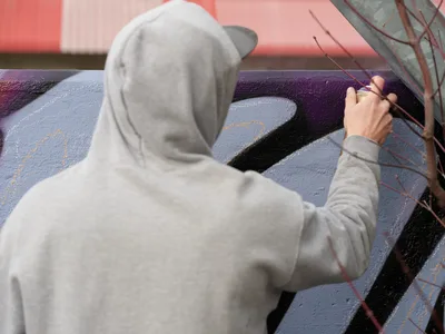 Travis Fields, known as Campographic, works intently on a vibrant mural at the Clay Creative Building in Portland's Hosford-Abernethy neighborhood. Dressed in a light gray hoodie, the artist's focused gestures guide crimson details across bold purple and blue geometric forms. The afternoon light catches the textured spray paint surface as creative energy transforms the urban canvas.
