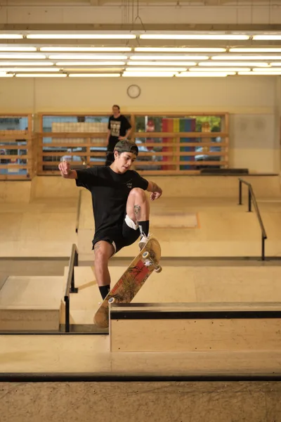 Professional skateboarder Christiana Means executes a precise frontside boardslide on a wooden ledge at Stronger Skatepark in Milwaukie, Oregon. The indoor facility's warm fluorescent lighting bathes the scene in amber tones, while geometric wooden ramps and rails create angular patterns throughout the background. A second skater observes from the upper level, adding depth to this moment of athletic focus and technical precision.