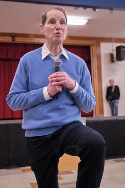 Senator Ron Wyden leans forward intently while speaking into a handheld microphone at a town hall meeting in Robert Gray Middle School's gymnasium in Portland, Oregon. The senior politician, dressed in a powder blue sweater over a crisp white collared shirt, commands attention from his seated position on the gymnasium's bleachers. Fluorescent lighting casts an even glow across the institutional setting, where red curtains frame wooden doors in the background and a lone attendee waits in the distance, creating an intimate yet formal atmosphere for civic engagement.