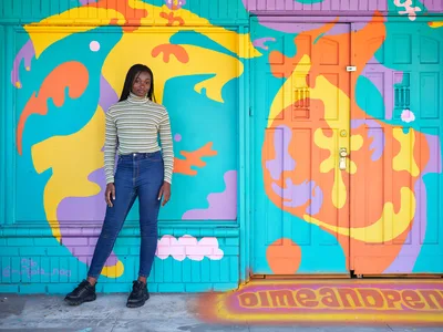 Naomi Likayi stands confidently beside her completed street mural on Northeast 15th Avenue in Portland's Alberta district, her signature visible in the corner of the psychedelic artwork. The young Black artist wears a striped turtleneck and jeans against the explosion of turquoise, orange, yellow and purple abstract forms that transform the building's facade. Warm afternoon light illuminates both the artist and her kaleidoscopic creation, capturing a moment of artistic completion in one of Portland's most vibrant neighborhoods.