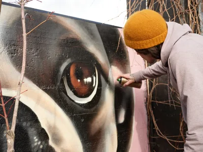 Muralist Wetiko meticulously applies spray paint to a striking hyperrealistic eye mural on an exterior wall in Portland's Hosford-Abernethy neighborhood. Wearing a golden yellow beanie and pale hoodie, the artist works with focused precision on the luminous amber iris, surrounded by detailed eyelashes and photorealistic skin tones. The intimate moment captures the transformation of urban space through art, with dried vegetation creating a natural frame around the creative process.