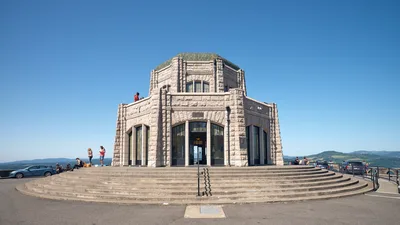 The iconic Vista House stands majestically atop Crown Point, its weathered stone facade gleaming under brilliant blue skies. Visitors gather on the circular stone steps and observation deck, drawn by the panoramic views of the Columbia River Gorge stretching toward distant forested hills. The Art Nouveau monument's distinctive octagonal architecture creates a striking silhouette against the endless Oregon sky, while tourists capture memories and explore this beloved Pacific Northwest landmark.