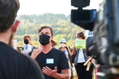Michael Gaskill, wearing a black face mask and v-neck shirt with a blue name tag, speaks earnestly to a KOIN Channel 6 news crew during a climate change protest at Representative Kurt Schrader's Oregon City office. The interview unfolds against a backdrop of forested hills bathed in golden afternoon light, while fellow demonstrators holding signs gather in the soft-focused background. The composition captures the intersection of grassroots activism and media coverage in the tree-lined McLoughlin Promenade setting.