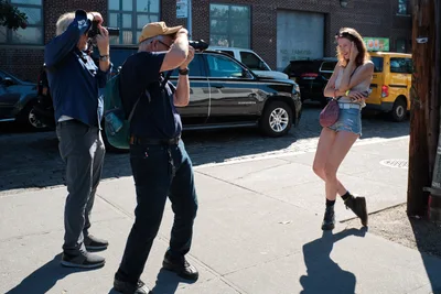 Two photographers work in golden hour light along a cobblestone street in DUMBO, Brooklyn, their telephoto lenses trained on a young woman posing against a wooden post. The subject, dressed in denim shorts and combat boots with a burgundy bag, strikes a casual pose while warm sunlight creates dramatic shadows across the industrial brick facade behind her. A black Chevrolet Suburban and yellow taxi provide urban context to this spontaneous street photography session in Emily Warren Roebling Plaza.