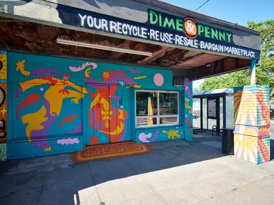 A vibrant turquoise storefront adorns the Dime A Penny recycle shop on Northeast 15th Avenue in Portland's Alberta district. The building bursts with psychedelic murals featuring whimsical creatures, abstract forms, and bold geometric patterns in electric oranges, purples, and yellows against the weathered wooden overhang. Afternoon sunlight bathes the concrete sidewalk, casting dramatic shadows that emphasize the contrast between the exuberant street art and the utilitarian urban architecture of this Northeast Portland neighborhood.
