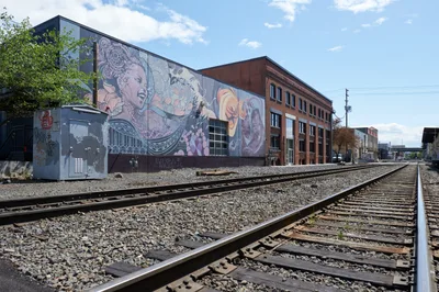 Steel railway tracks stretch into the distance through Portland's industrial Buckman neighborhood, flanked by weathered brick buildings adorned with vibrant street murals. The elaborate artwork depicts flowing figures and ornate patterns in rich purples, oranges, and earth tones, creating a striking contrast against the utilitarian infrastructure. Afternoon light illuminates the scene under a partly cloudy sky, while fresh spring foliage frames the urban tableau where transportation corridors intersect with public art.