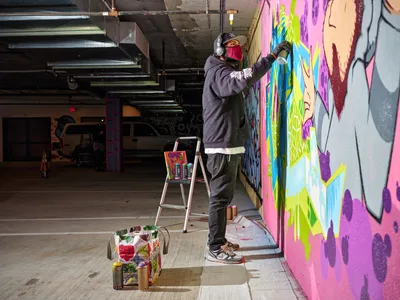 In the industrial depths of Clay Creative's Southeast Portland parking garage, a masked muralist applies brilliant strokes of magenta, lime green, and azure to transform concrete walls into living art. The artist, wearing headphones and a crimson face covering, stands atop a metal stepladder surrounded by scattered spray cans and paint containers. Exposed ductwork and fluorescent lighting cast harsh shadows across the cavernous space, while the emerging mural bursts with color against the stark urban infrastructure.