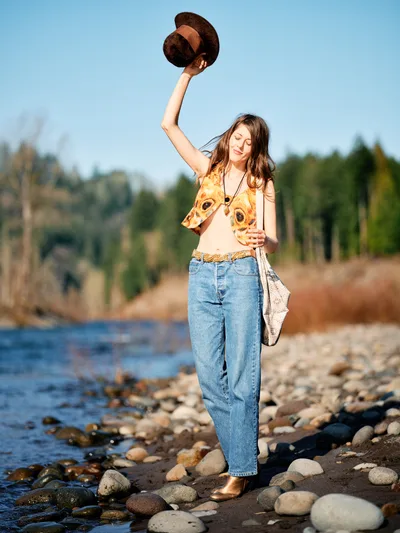 A young woman in vintage-inspired attire raises her wide-brimmed hat triumphantly along the rocky shoreline of Sandy River in Oregon's Floodplain Trail. The golden hour light illuminates her sunflower-print vest and creates a warm, nostalgic atmosphere as gentle waters lap against the smooth river stones. Her joyful gesture and closed eyes convey a moment of pure liberation and connection with nature.