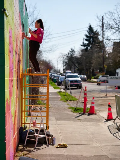 Multidisciplinary artist Vilatose stands atop scaffolding, spray paint in hand, adding bold strokes to a rainbow-hued mural cascading down a brick wall along Northeast Alberta Street. The afternoon light illuminates her focused expression and magenta shirt as she works against the backdrop of Portland's Alberta Arts District, where orange traffic cones mark the creative transformation in progress. Paint supplies and a stepladder scatter the sidewalk below, evidence of the urban canvas taking shape.
