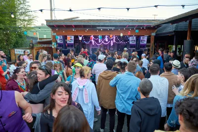 A vibrant crowd fills the outdoor patio of White Owl Social Club in Portland, Oregon, during the season opener of Your Sunday Best, a popular summer day party. Revelers in colorful costumes and casual attire dance beneath string lights and festive decorations, with the venue's covered stage area glowing with purple lighting in the background. The late afternoon atmosphere captures the essence of Portland's eclectic music scene, as dozens of party-goers embrace the celebratory mood of the season's first outdoor dance gathering.
