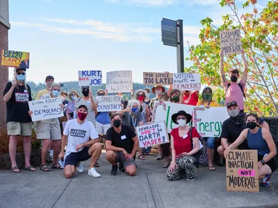 Masked protesters gather in a carefully arranged group portrait on the sidewalk outside Representative Kurt Schrader's Oregon City office, their handmade signs demanding climate action and voting rights creating a mosaic of grassroots activism. The late afternoon light bathes the diverse crowd of approximately twenty participants, ranging from young adults to seniors, all unified in their call for clean energy policy and democratic reform. Their colorful protest signs, featuring messages like "Climate Inaction is NOT Moderate" and "Kurt, Back Joe," stand out against the backdrop of brick buildings and autumn foliage along McLoughlin Promenade.