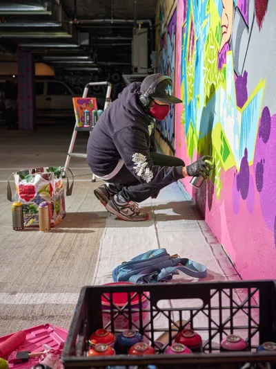 A masked street artist crouches intently beside a vibrant mural in progress, spray can in hand, within the industrial confines of Clay Creative's Southeast Portland parking garage. The scene captures the focused concentration of urban art creation, surrounded by crates of colorful spray paint and the raw concrete textures of the converted creative space. Brilliant neon pinks, electric blues, and lime greens cascade across the wall, contrasting sharply with the artist's dark hoodie and the garage's utilitarian atmosphere. The composition frames the intersection of industrial architecture and contemporary street art culture in Portland's creative district.