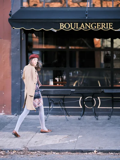 A young woman in a burgundy beret and camel coat strides past a French boulangerie on Northeast Alberta Street in Portland's Alberta district. Her outfit combines classic Parisian elements—the wool beret, tailored coat, and plaid skirt—with comfortable walking attire as she moves along the weathered sidewalk. The dark storefront with its elegant gold lettering creates an atmospheric backdrop, while natural light catches her mid-stride, suggesting a spontaneous street photography moment in this gentrified neighborhood.
