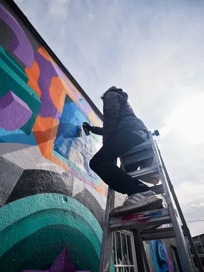 A bundled artist perches on aluminum scaffolding against a cloudy Pacific Northwest sky, spray painting an explosive kaleidoscope of geometric shapes across a building wall in Northeast Portland. The mural blooms with emerald greens, electric purples, burnt oranges, and azure blues, creating bold abstract forms that transform the urban landscape. The low-angle composition emphasizes the scale of the artistic endeavor while soft overcast light bathes the scene in muted tones typical of Portland's atmospheric conditions.