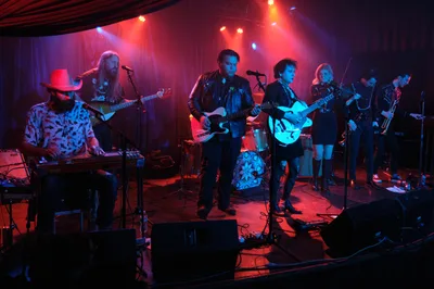 Bathed in crimson and azure stage lighting, six-piece band Roselit Bone delivers an intimate performance at the legendary Dante's venue in Portland, Oregon. The atmospheric lighting cuts through wisps of stage fog, illuminating the diverse ensemble of musicians wielding guitars, keyboards, and brass instruments. From left to right, a bearded keyboardist in a cowboy hat anchors the sound while electric guitars and horns create layers of melody across the small stage, their silhouettes dramatically carved by the moody concert illumination.
