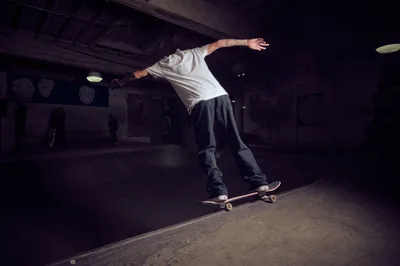 A tattooed skateboarder executes a fluid trick in the moody underground space of Commonwealth Skateboarding in Portland's Buckman neighborhood. The dimly lit basement venue creates dramatic shadows across weathered concrete, while exposed ceiling beams and vintage fixtures frame the athlete's precise movement. The skater's white shirt contrasts against the dark industrial interior, capturing the raw authenticity of Portland's skateboarding culture.