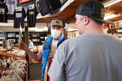 In the warm, wood-paneled interior of Music Millennium on East Burnside Street during Record Store Day 2022, a masked customer wearing headphones and a camouflage cap examines a vinyl record while conversing with another patron. The scene captures the tactile ritual of record browsing, with towering wooden bins filled with albums creating an intimate, nostalgic atmosphere under the store's fluorescent lighting.