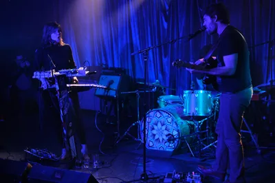 Experimental band Sun Blood Stories performs in the intimate confines of Dante's in downtown Portland during Rx Fest, bathed in deep cobalt stage lighting. The Boise-founded duo commands the small venue with synthesizers and drums, their silhouettes emerging from the moody blue atmosphere as theatrical curtains frame the performance. Electronic equipment and cables sprawl across the darkened stage, creating an authentic indie music scene captured in the heart of Portland's underground music culture.
