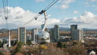 A sleek silver gondola car of the Portland Aerial Tram floats gracefully through autumn air, its reflective surface catching the soft overcast light above the South Waterfront district. The panoramic view reveals Portland's modern high-rise towers nestled between the Willamette River and rolling hills, while bare-branched trees in the foreground signal late fall's arrival. The composition captures both the engineering marvel of the tram system and the urban landscape it serves, connecting OHSU's Marquam Hill campus to the bustling waterfront below.