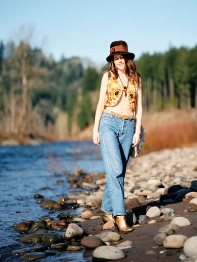 A young woman with flowing brown hair stands confidently on the rocky shoreline of Oregon's Floodplain Trail, her vintage-inspired outfit creating striking contrast against the natural landscape. Golden hour light illuminates her brown fedora hat, graphic vest, and high-waisted jeans as she poses beside the gentle waters. The composition captures her relaxed stance among smooth river stones, with the soft-focus forest backdrop emphasizing the connection between fashion and nature. Her warm smile and bohemian styling evoke a sense of free-spirited adventure in this serene riparian setting.
