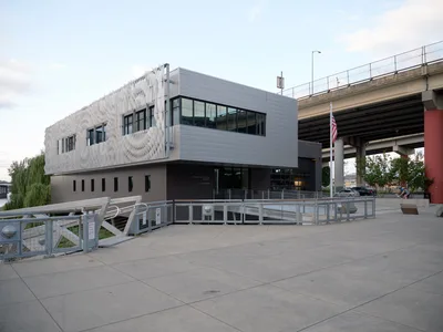 Portland Fire & Rescue Station 21 emerges as a striking example of contemporary public architecture beneath the Interstate 84 overpass in the Central Eastside district. The building's sculptural white facade features an undulating parametric pattern that contrasts dramatically with the corrugated metal upper level and industrial concrete surroundings. Captured during the golden hour transition, the structure's modern design philosophy transforms utilitarian emergency services architecture into an urban landmark that bridges infrastructure and community.