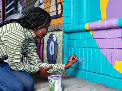 Naomi Likayi kneels beside her vibrant street mural in Portland's Alberta Arts District, carefully painting her signature in purple letters on a brilliant turquoise section of the wall. Her intricate braided hair catches the afternoon light as she concentrates on her work, wearing a striped long-sleeve shirt and jeans. Behind her, the colorful geometric mural explodes across the brick wall in bold strokes of purple, yellow, and cyan, transforming the urban corner of Northeast 15th and Killingsworth into a canvas of community expression.