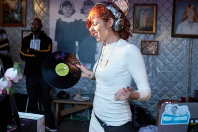 DJ Doctor Dakar commands the booth at White Owl Social Club, her flame-red hair catching golden light as she grips a vintage vinyl record with infectious enthusiasm. The intimate Portland venue pulses with afternoon energy, its walls adorned with eclectic artwork creating a bohemian backdrop. Her pure joy radiates through professional headphones and crisp white attire, embodying the authentic spirit of vinyl culture in the Pacific Northwest music scene.