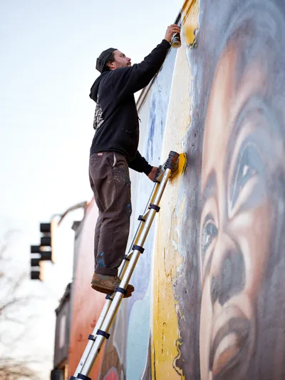 Travis Fields, known as campographic, stretches upward on an aluminum ladder to apply paint to a vibrant mural depicting poet Amanda Gorman. The street artist works against weathered concrete in Portland's Alberta Arts District, his dark hoodie and paint-stained work pants capturing the dedication of public art creation. Soft afternoon light illuminates the emerging portrait, revealing layers of blue, yellow, and flesh tones that bring the celebrated poet's likeness to life on the urban canvas.
